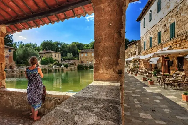 A woman admires the scenic thermal pool of Bagno Vignoni, Tuscany, surrounded by historic stone buildings and charming outdoor cafés.