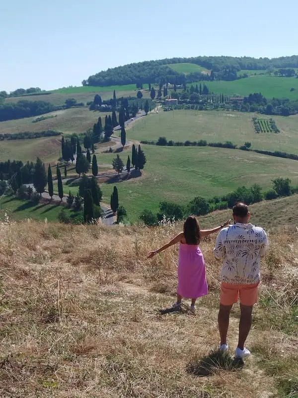 Couple enjoying a scenic view of the Tuscan countryside: Two people standing on a dry hillside overlooking rolling green fields and a winding road lined with cypress trees. Ideal for promoting scenic tours and romantic getaways in Tuscany.
