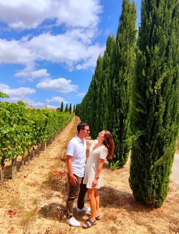 A couple enjoying a private Tuscany tour, standing between lush vineyards and towering cypress trees under a bright blue sky, capturing the essence of an authentic Italian countryside experience.