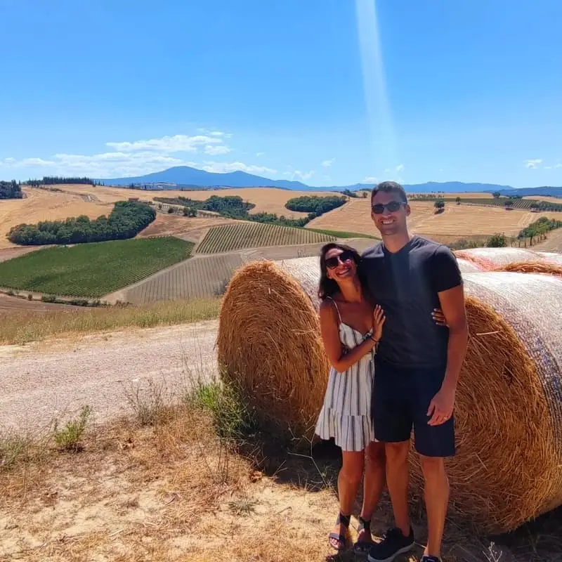 Couple posing with hay bales in Tuscan landscape: A man and woman standing next to large hay bales, with vineyards and rolling hills in the background under a blue sky. Ideal for promoting scenic tours and romantic getaways in Tuscany.