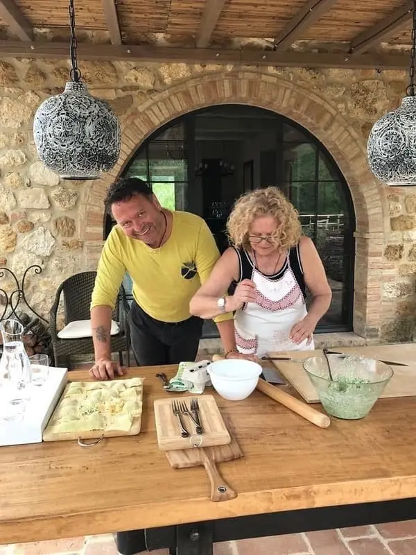 Fabrizio and his mother teaching a pasta-making class in Tuscany: Fabrizio and his mother demonstrate pasta preparation at a wooden table with ingredients, in a Tuscan outdoor setting with stone arches. Ideal for promoting cooking classes and culinary experiences in Tuscany with Fabrizio.