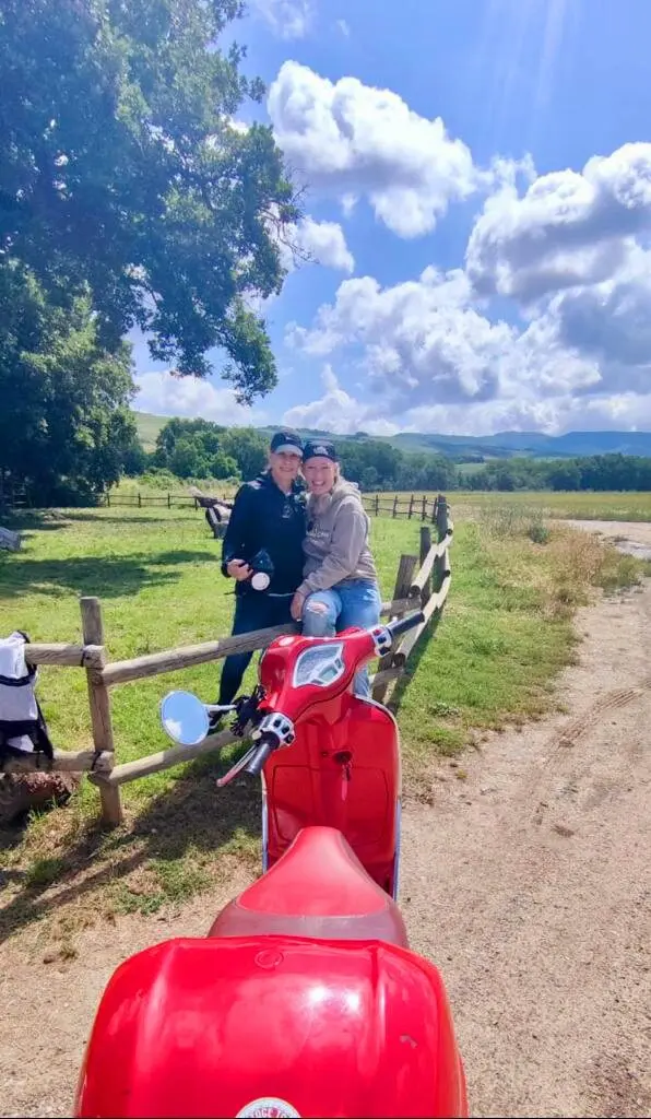 Ciao Fabry Tours: Two people posing with a red Vespa scooter on a Tuscan countryside tour. Scenic landscape with green fields, wooden fence, and blue sky with clouds. Ideal for promoting Vespa tours in Tuscany and outdoor travel experiences.