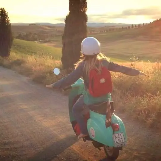 Woman riding a Vespa scooter in the Tuscan countryside: A woman with a white helmet and red backpack rides a teal Vespa on a dirt road, arms outstretched, with the text 'X4T LBT' on the license plate. Scenic landscape with rolling hills and cypress trees in the background. Ideal for promoting Vespa tours and scenic drives in Tuscany.