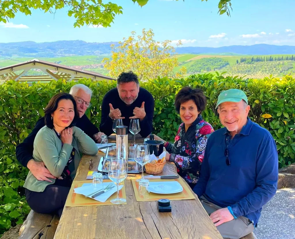Fabrizio with two couples enjoying a wine tour meal in Tuscany: Fabrizio poses with two couples at a wooden table with wine glasses and plates, with a scenic Tuscan landscape in the background. Ideal for promoting wine tours and culinary experiences in Tuscany with Fabrizio.