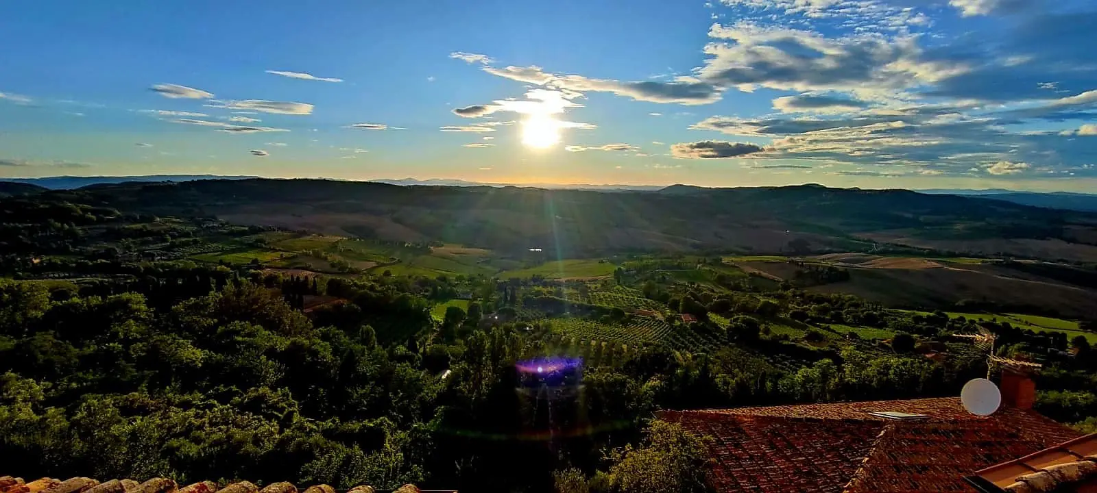 Sunset over the Tuscan countryside: Panoramic view of rolling hills and valleys with a bright sun setting against a blue sky with clouds. Ideal for promoting scenic tours and landscape photography in Tuscany.