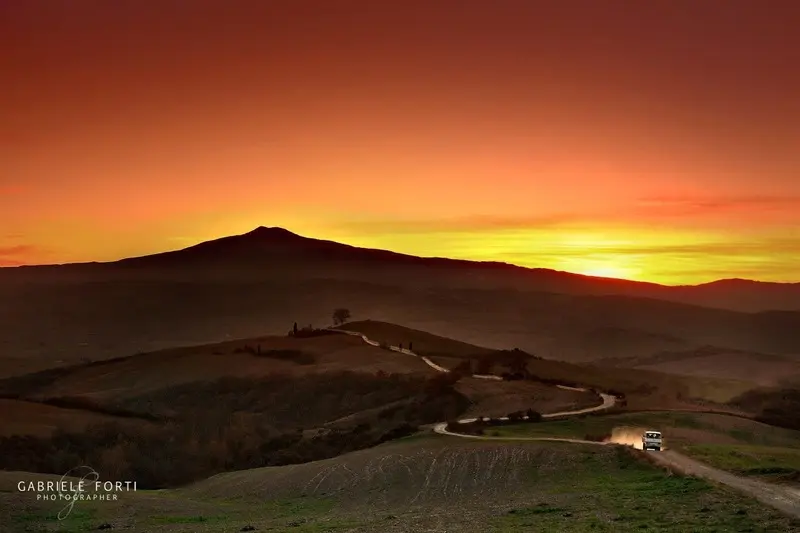 Sunset over Mount Amiata, Tuscany: A scenic view of rolling hills with a winding road leading towards Mount Amiata, silhouetted against a vibrant orange and yellow sunset. A small vehicle is visible on the road. Photo by Gabriele Forti. Ideal for promoting scenic drives and landscape photography in Tuscany.