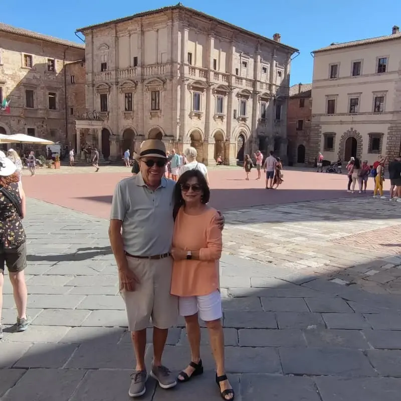 Couple in Piazza Grande, Montepulciano, Italy: Two tourists posing for a photo in the historic Piazza Grande, with the Palazzo Comunale in the background. Ideal for promoting Montepulciano tours and sightseeing in Tuscany.