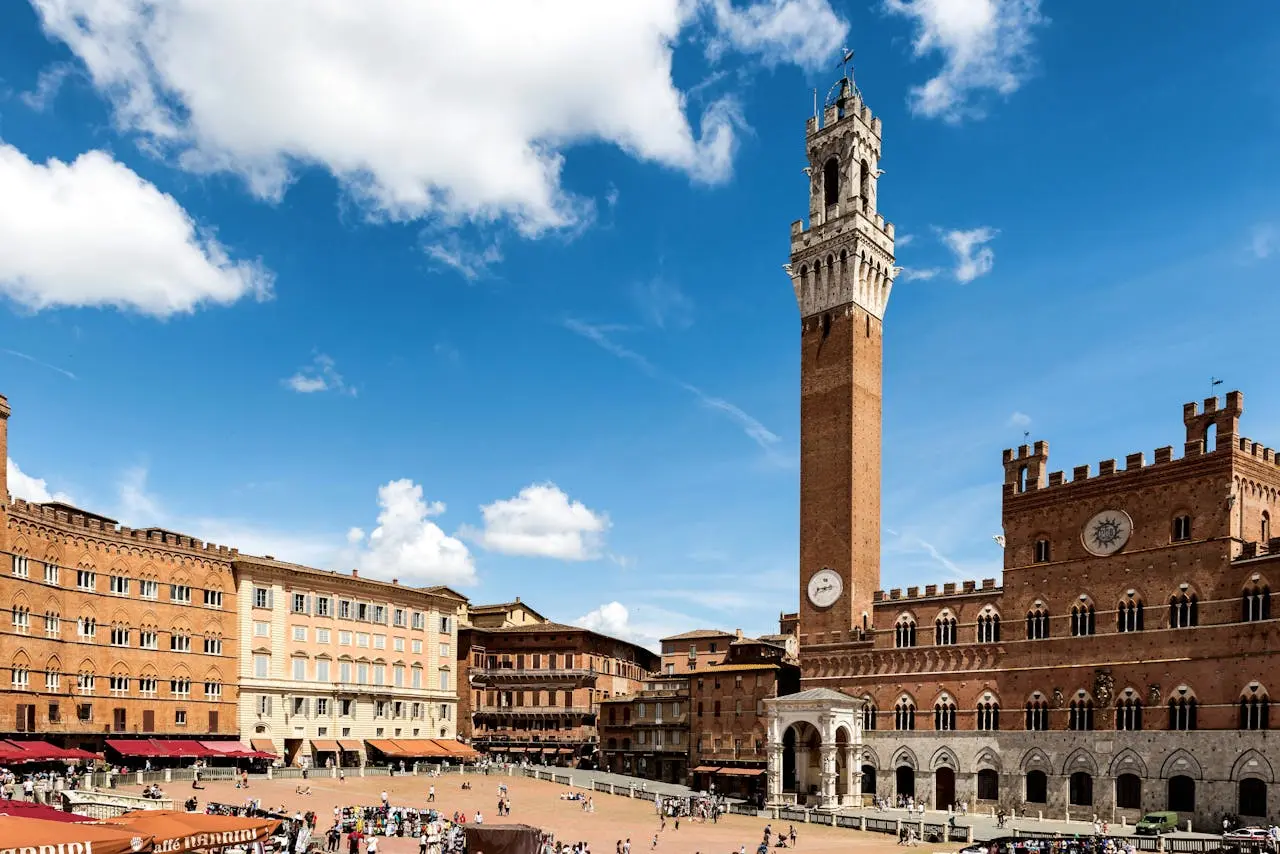 Piazza del Campo, Siena, Tuscany, Italy: Panoramic view of the historic square with the Palazzo Pubblico and Torre del Mangia. Tourists gather in the square under a bright blue sky with white clouds. Ideal for promoting Siena sightseeing and cultural tours.