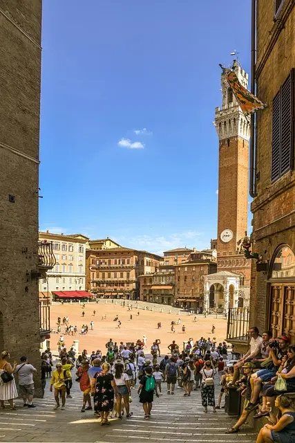 Piazza del Campo, Siena, Tuscany, Italy: A bustling square with tourists walking towards the Palazzo Pubblico and Torre del Mangia. Historic Italian architecture and a vibrant atmosphere. Ideal for promoting Siena tours and cultural experiences in Tuscany.