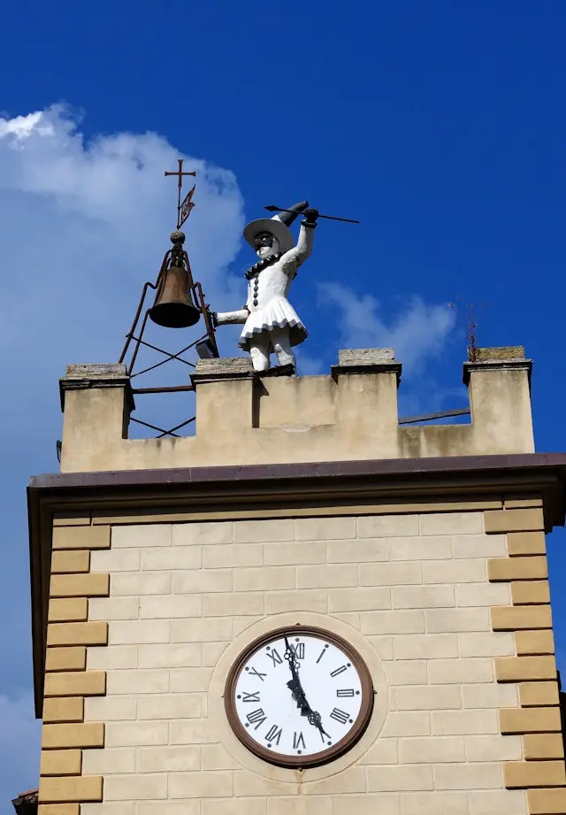 Pulcinella Tower in Montepulciano, Tuscany, Italy: A clock tower featuring a Pulcinella figure striking a bell, with a bright blue sky and white clouds in the background. Architectural landmark and tourist attraction in Montepulciano.