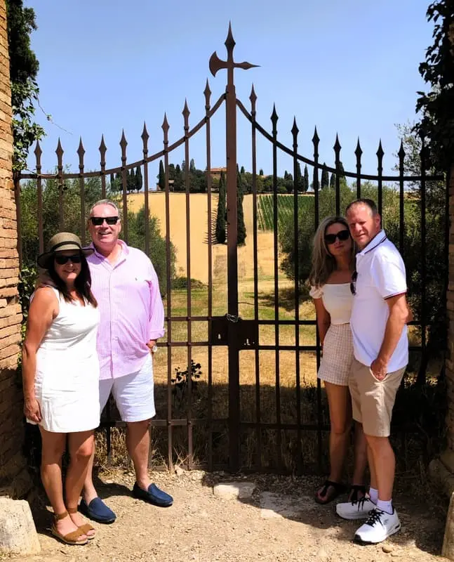 Group tour in Tuscany: Four people posing in front of an ornate iron gate with a scenic Tuscan landscape in the background. Ideal for promoting guided tours and travel experiences in Tuscany.