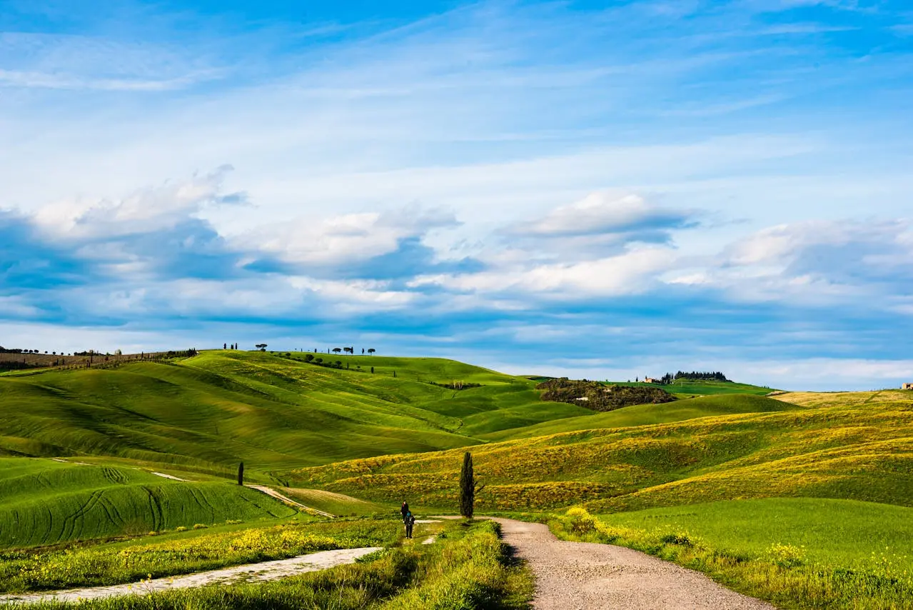 Val d'Orcia landscape, Tuscany: A scenic view of rolling green hills, a dirt road, and a bright blue sky with white clouds. Ideal for promoting scenic drives and landscape photography in the Val d'Orcia region of Tuscany.