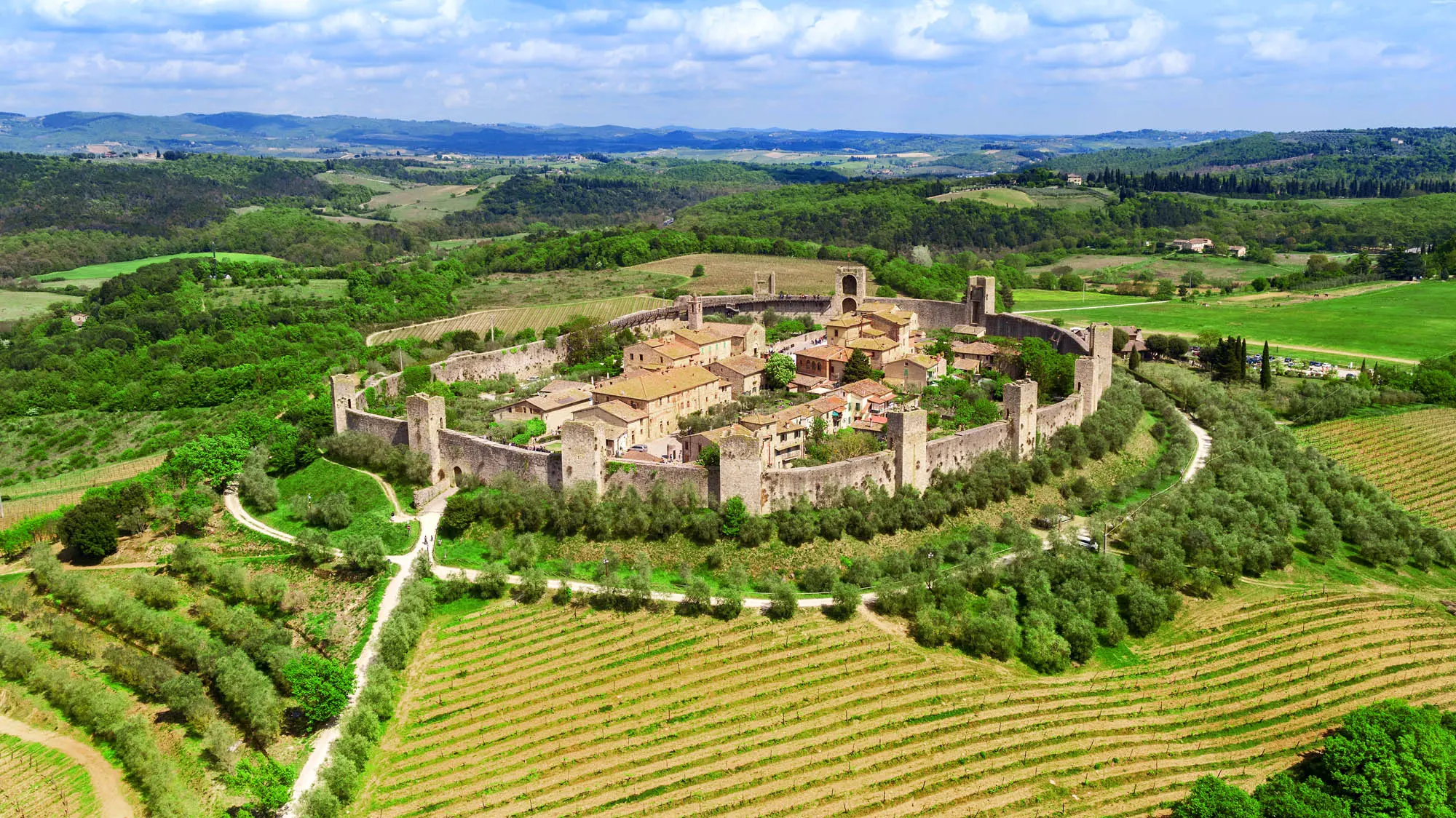 Aerial view of Monteriggioni, a walled medieval town in Tuscany, Italy, surrounded by green rolling hills and vineyards. A circular stone wall encloses the town with visible towers, showcasing historical architecture. Ideal for Tuscany tours and historical sightseeing.