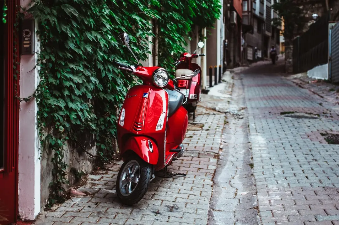 Red Vespa scooters parked on a cobblestone street in an urban setting, possibly for a Vespa tour. Lush green vines on a building wall add a touch of nature. Ideal for promoting city Vespa tours and Italian urban travel experiences.