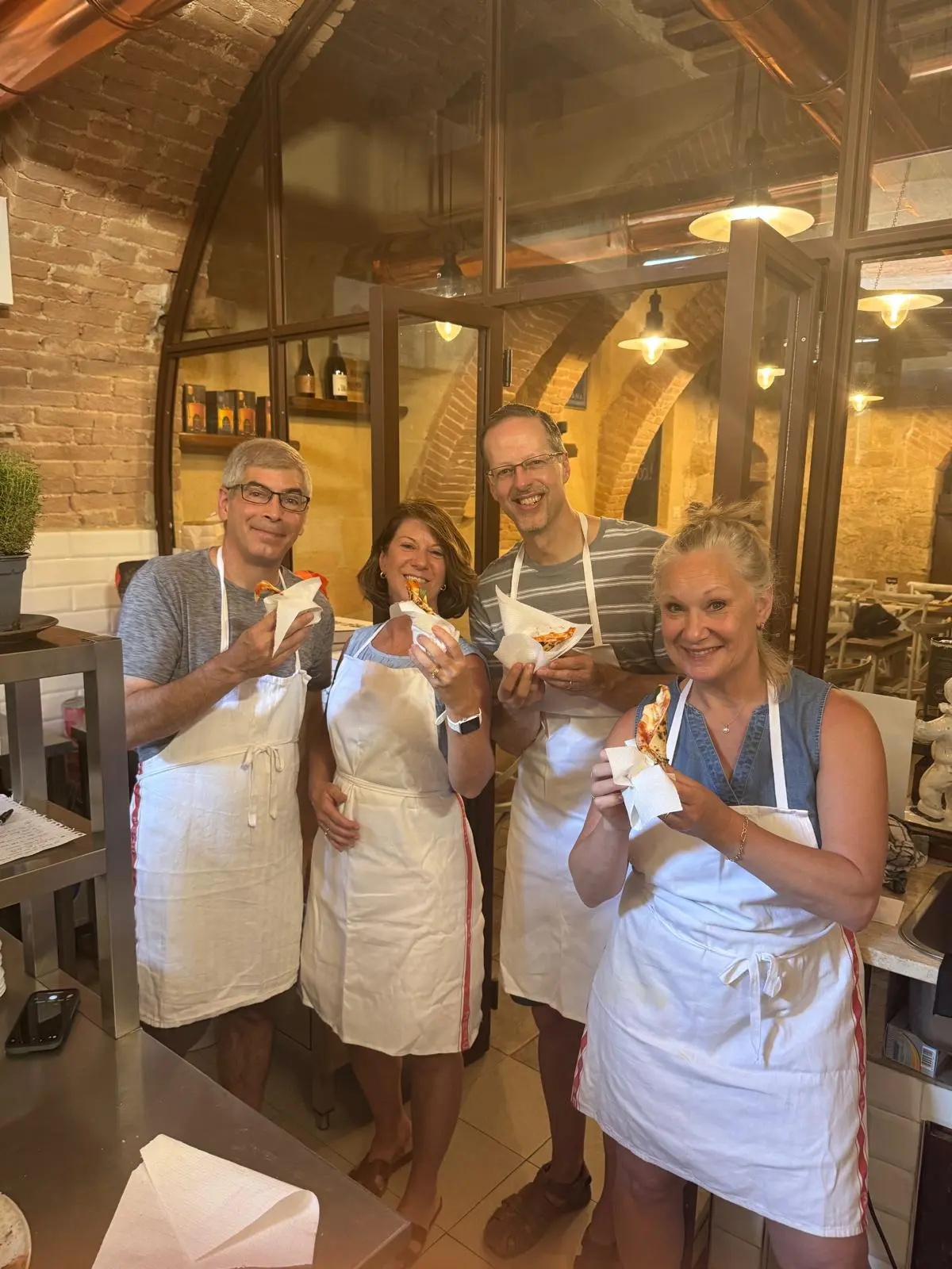 Four smiling adults, wearing white aprons, proudly holding and eating freshly made pizza slices in a rustic kitchen setting, clearly enjoying their pizza making class in Montepulciano, Tuscany. This image captures the authentic culinary experience offered during a cooking class in Italy.
